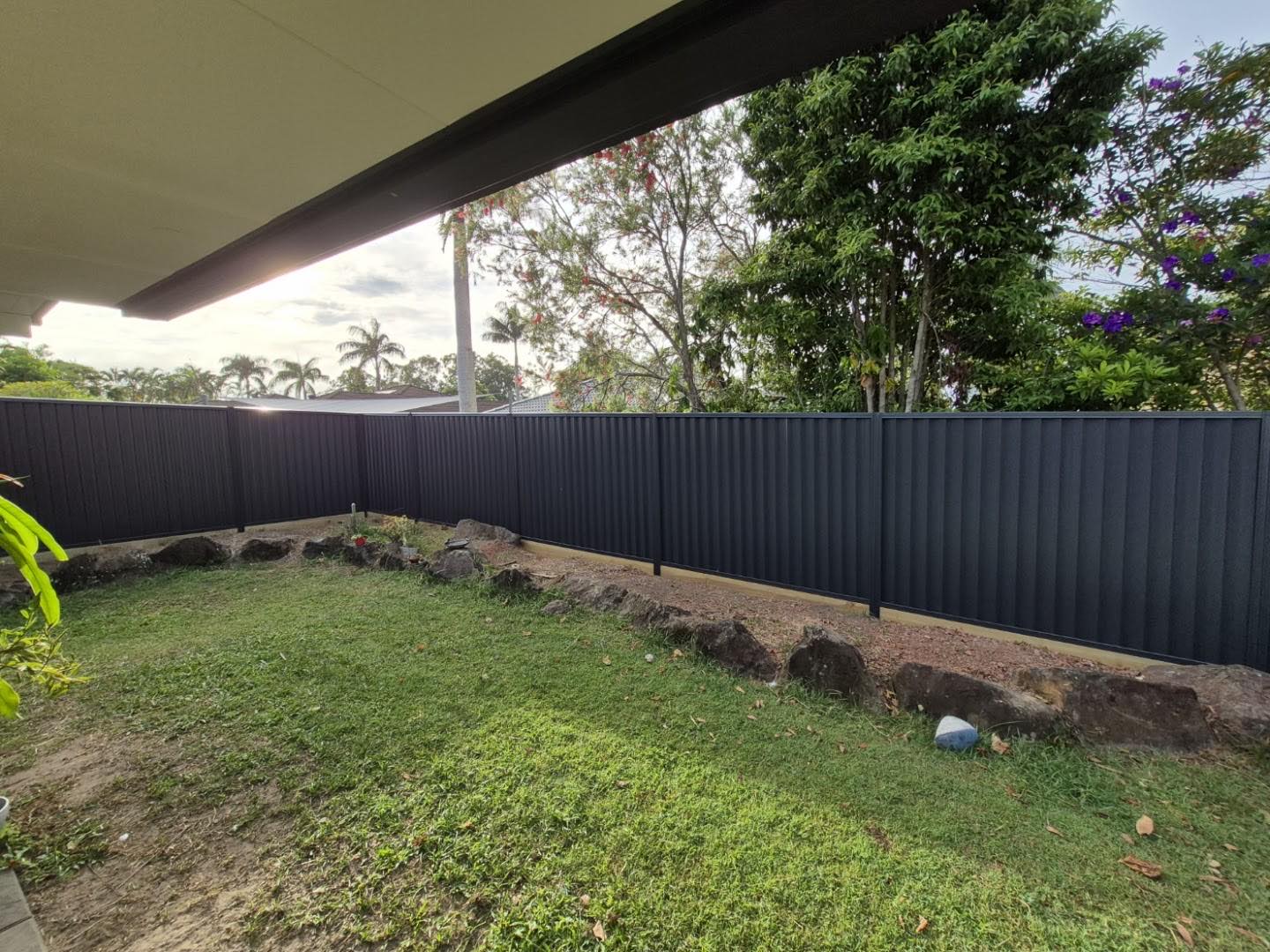 A dark Colorbond fence running along a lawn under the shade of a large tree