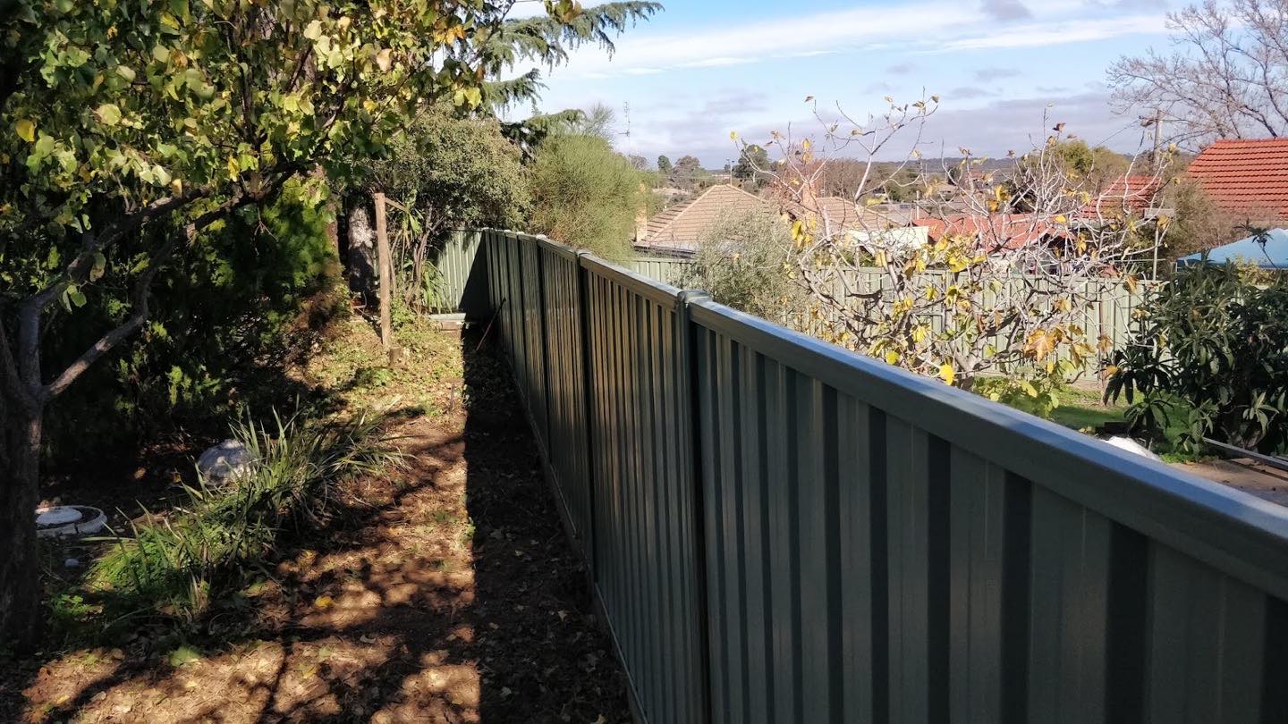 A dark Colorbond fence running beside a mulched garden bed under trees