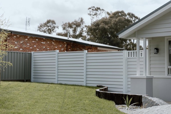 A cream slatted fence running past a brick home and a garden bed